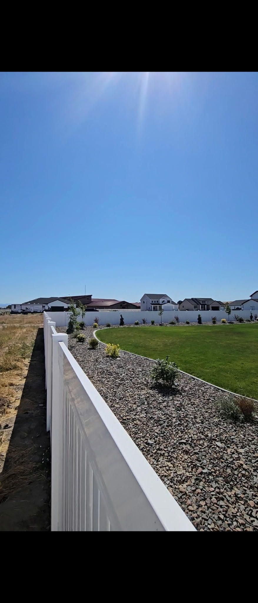 White fence along gravel path beside green field under a bright blue sky with distant houses