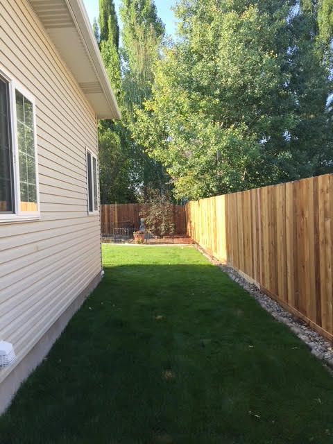 Narrow grassy side yard between a beige house and wooden fence, with trees in the background.