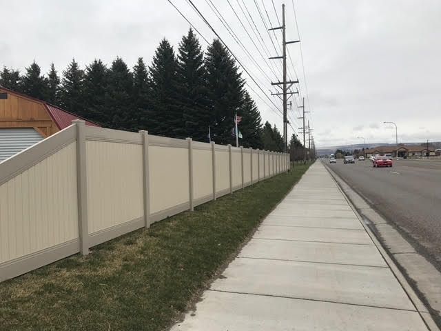 Sidewalk beside a beige fence along a road, with power lines and trees in the background.