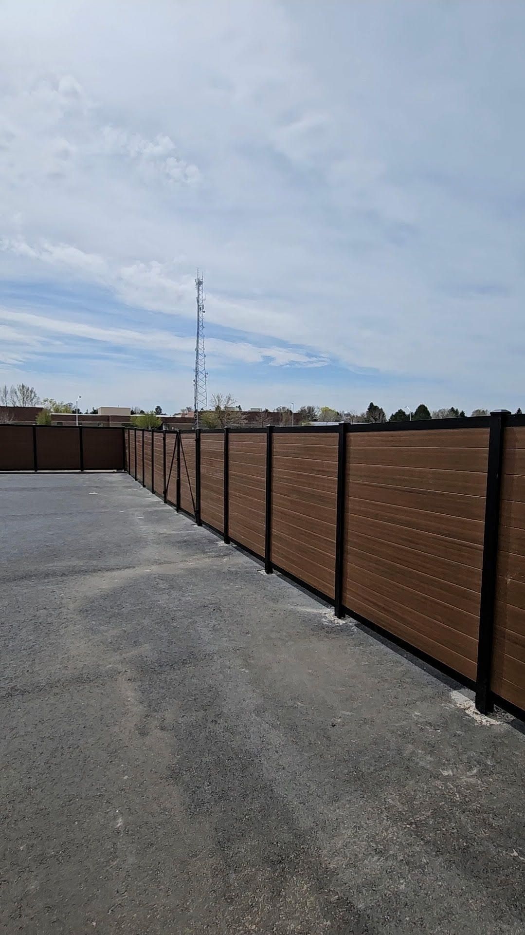 Wide paved lot beside a long brown fence under a cloudy sky, with a tall utility tower in the distance