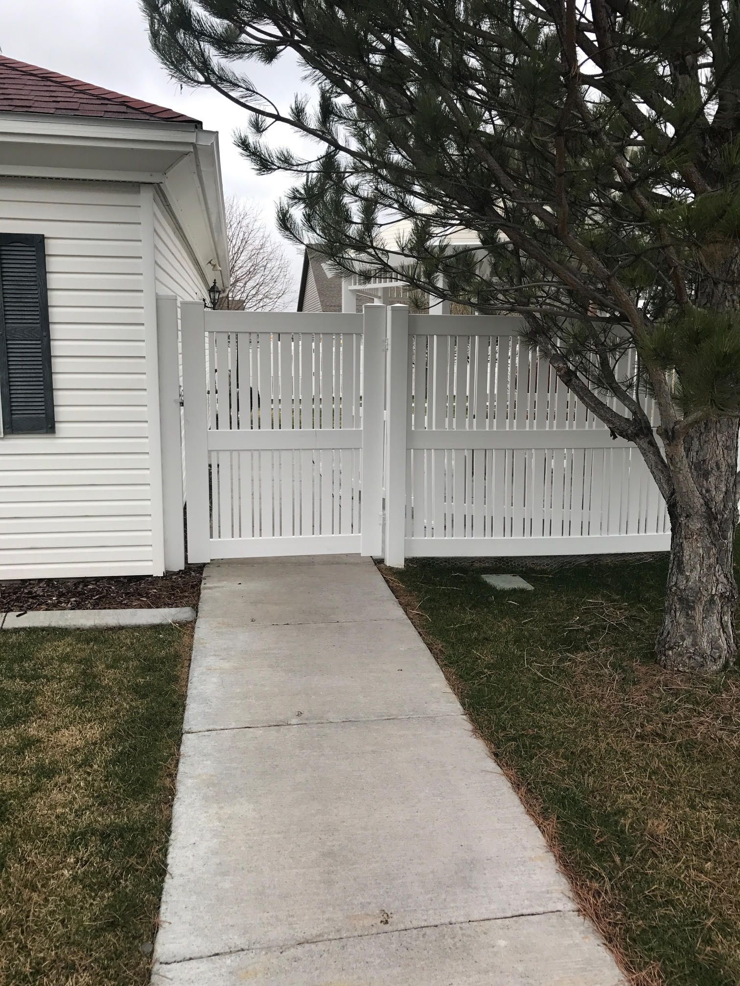 Concrete walkway leading to a white fence gate beside a house and tree.