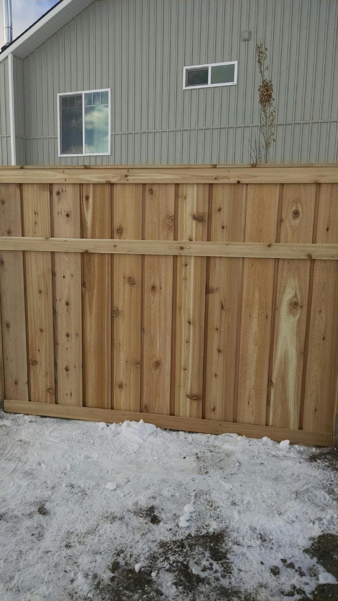 Wooden privacy fence with snow on the ground beside a gray house wall and windows