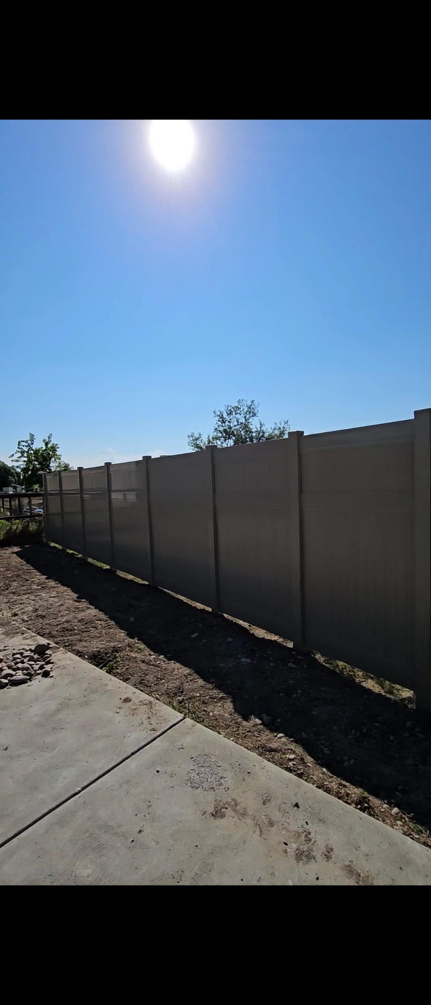 Dark metal fence with angled slats along a concrete wall beside trees and red flowers