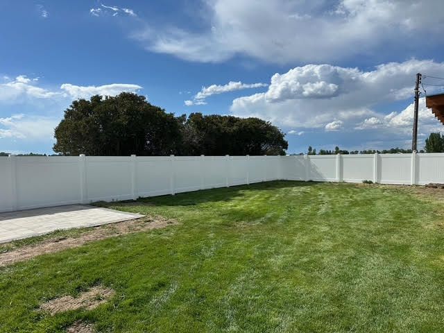Backyard with green lawn, white fence, concrete patio, and blue sky with clouds