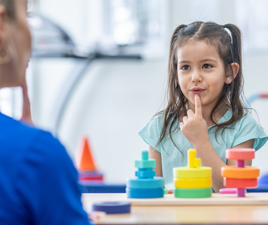 Child playing a matching game, touching her lips, with colorful stacking toys on a table in a classroom