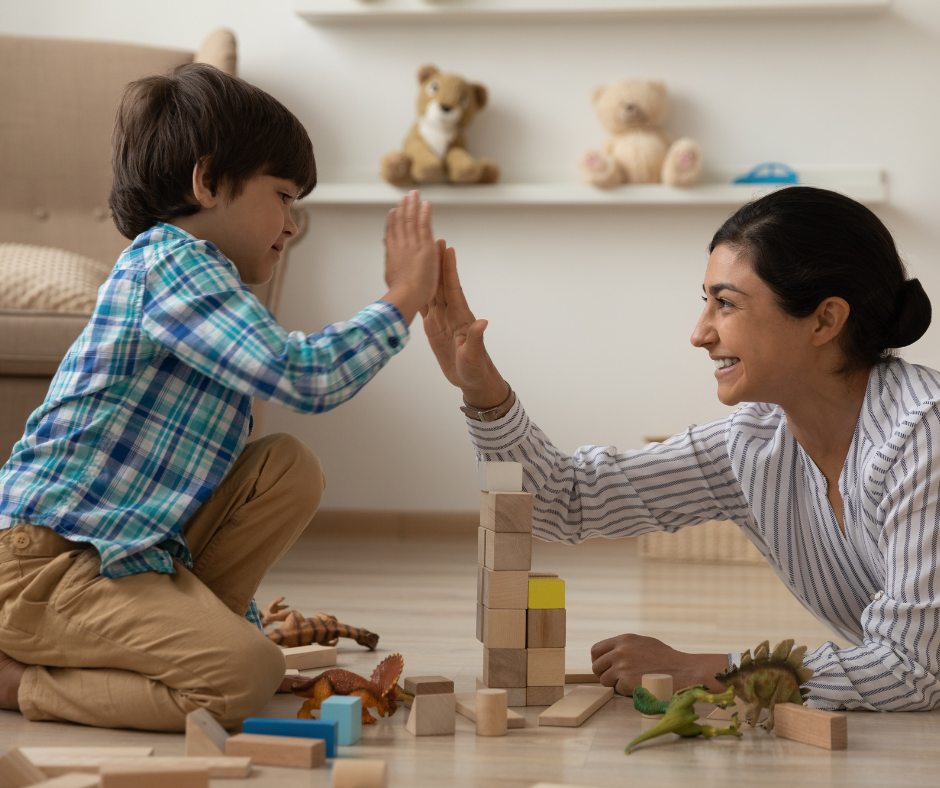 Child and woman smiling, giving a high-five while playing with wooden blocks on the floor.