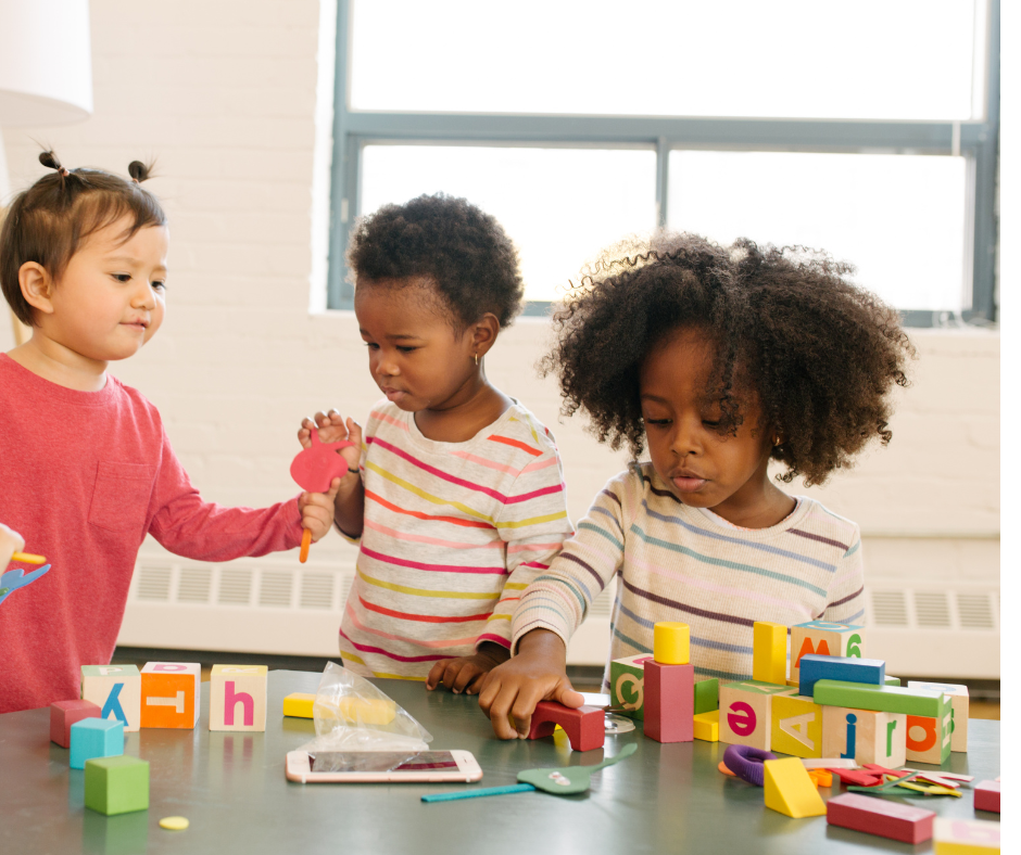 Three children playing with colorful blocks at a table in a bright classroom