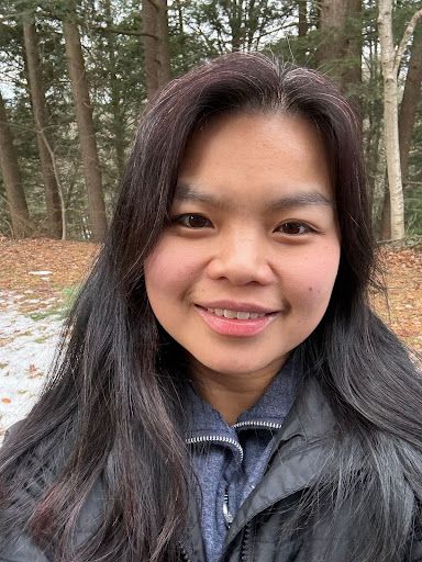 Smiling person in a gray jacket standing outdoors among tall trees in a wooded area
