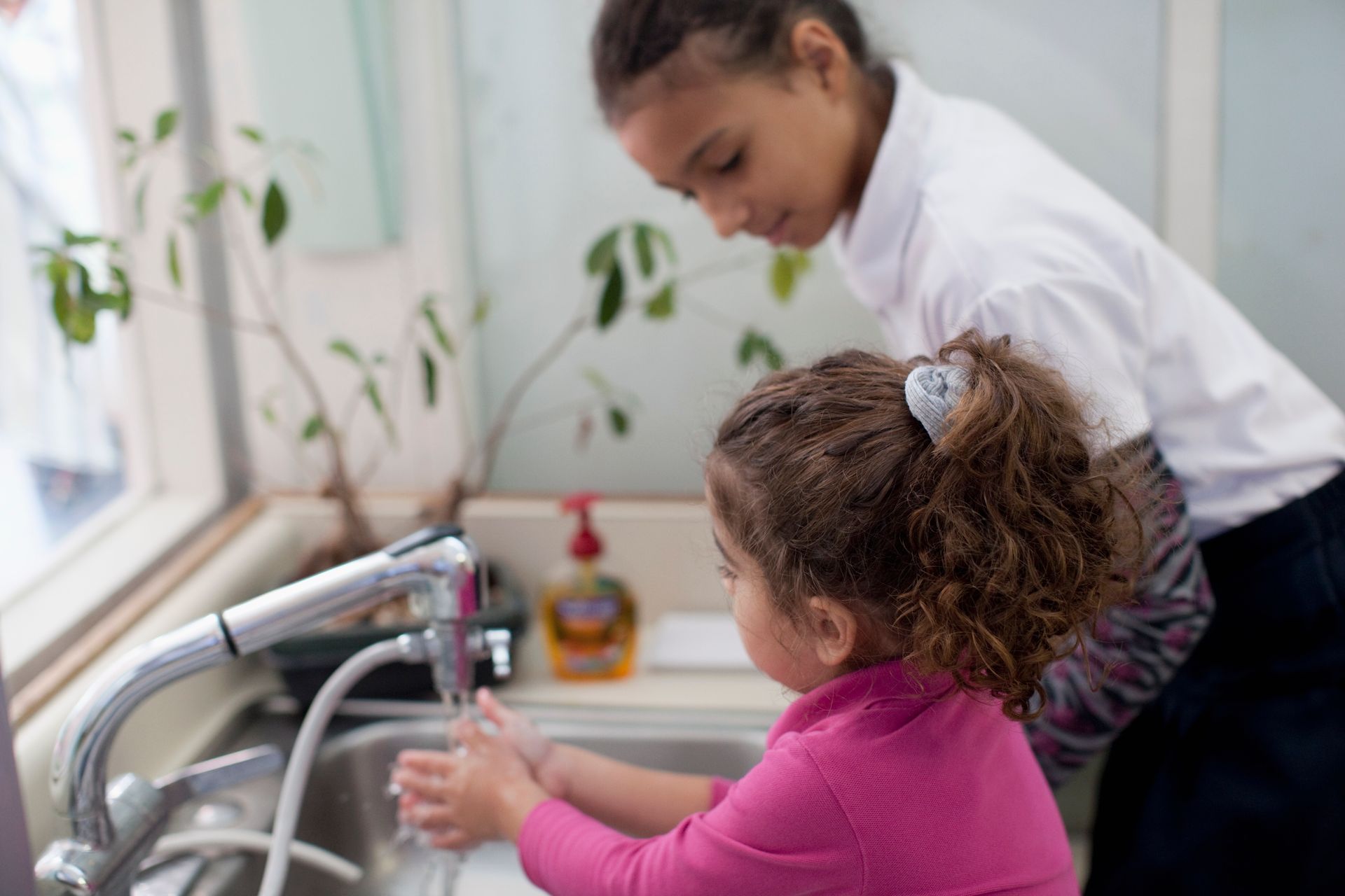 Two children washing hands at a sink, one helping the other with soap and water.