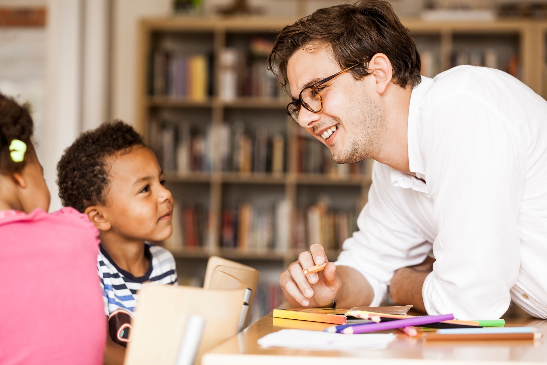 Teacher smiling and talking with children at a classroom table with books and papers