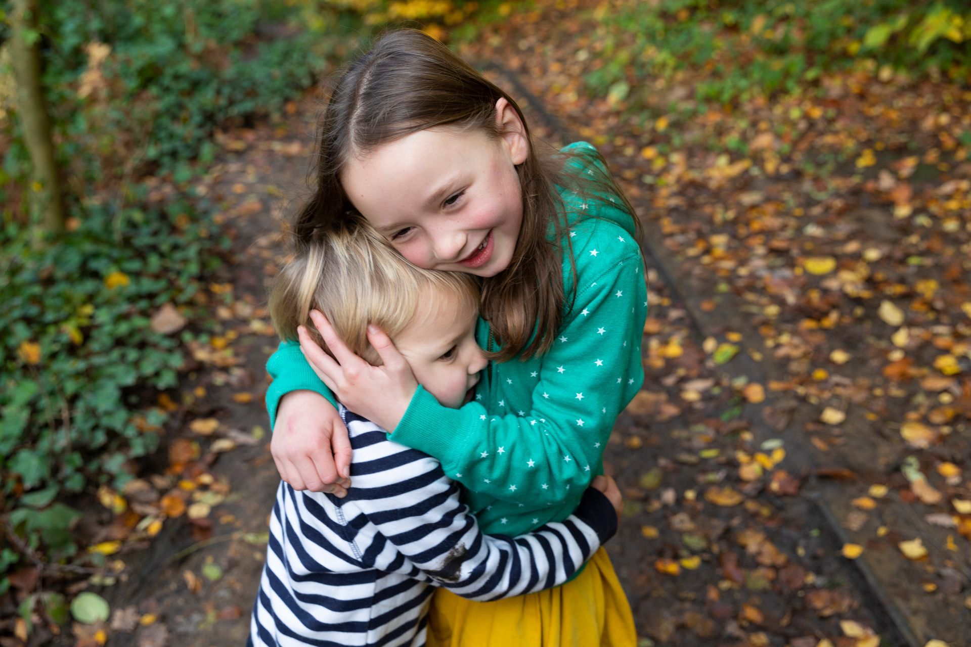 Two children hugging on a leafy autumn path, smiling warmly.