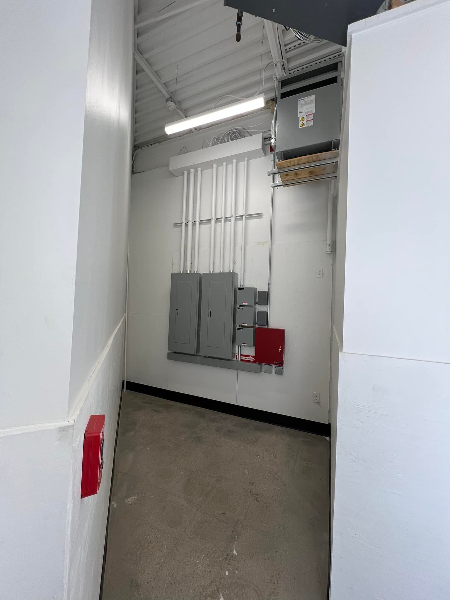 Narrow room with electrical panels on the back wall. White walls, grey floor. A fire alarm is visible.