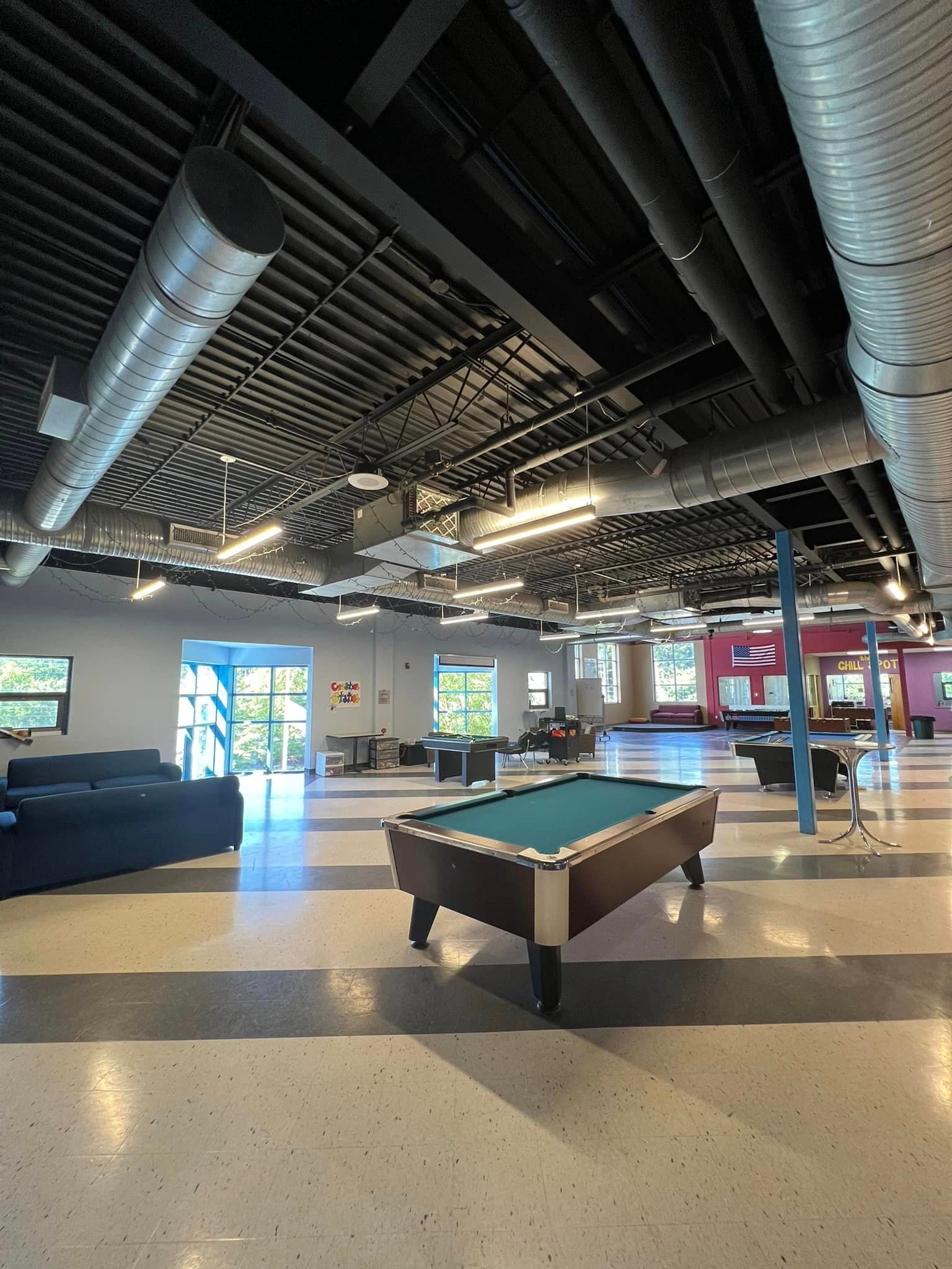 Large room with a pool table, couches, and exposed ceiling ductwork. Windows and seating in the background.