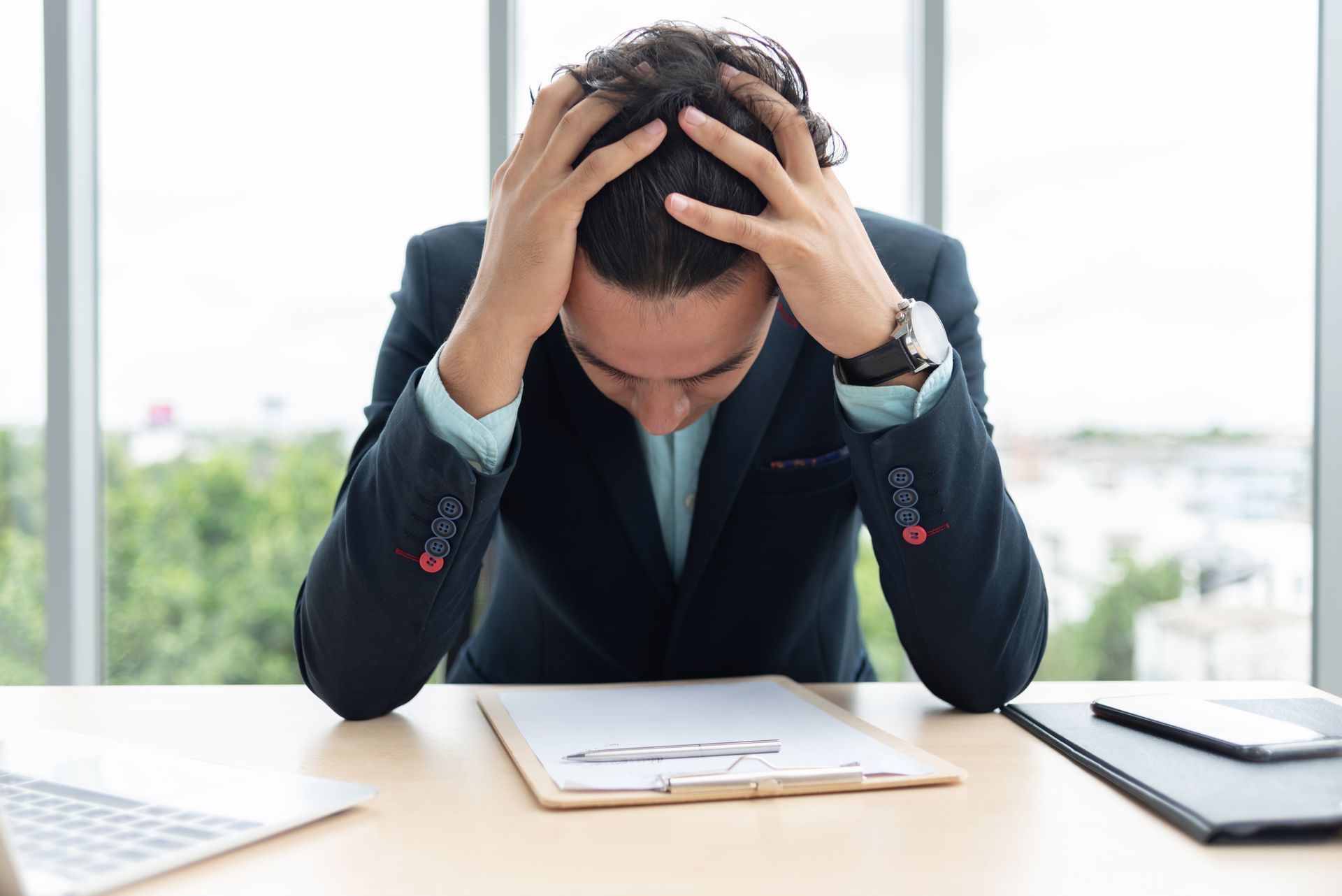 Man in suit with hands on head, looking down at paperwork, stressed in office.