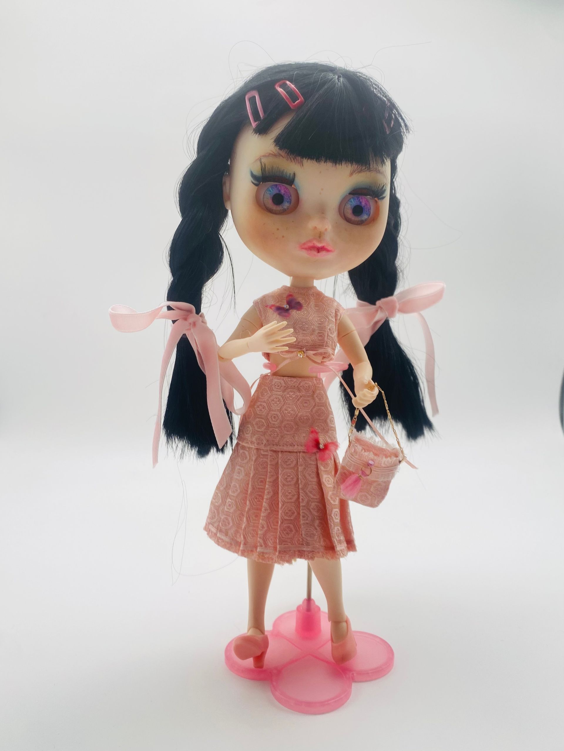 Pop in roze outfit, met zwarte gevlochten staartjes en roze accessoires, staand op een roze voetstuk.