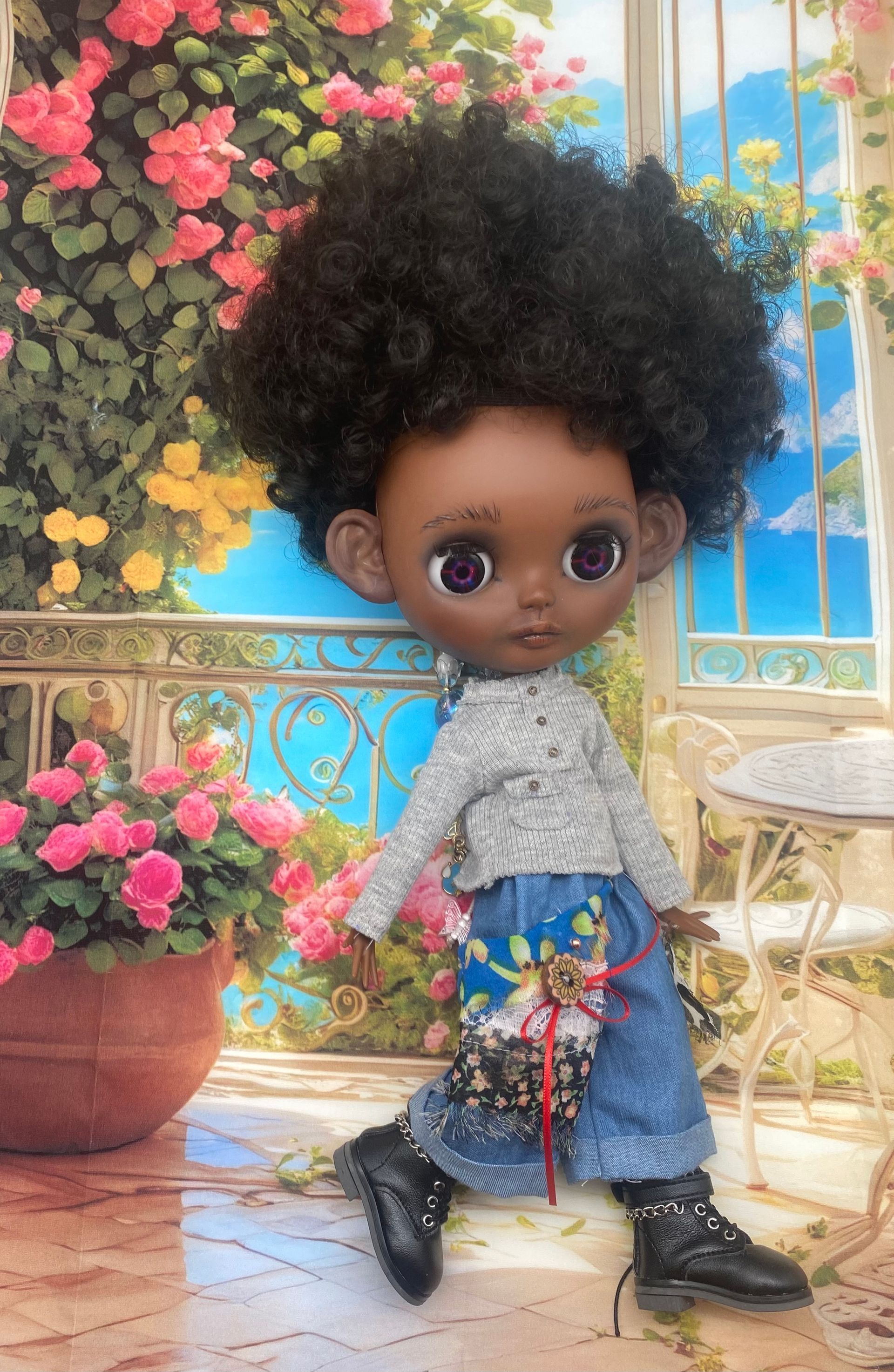 Een pop met een donkere huid, een grote afro, gekleed in een trui, jeans en laarzen, poseert voor een tuin vol bloemen.