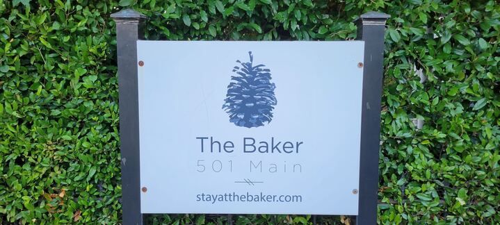 A sign for the baker 501 main is sitting in front of a hedge.