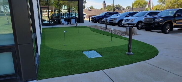 A small putting green in front of a building with cars parked in the background.