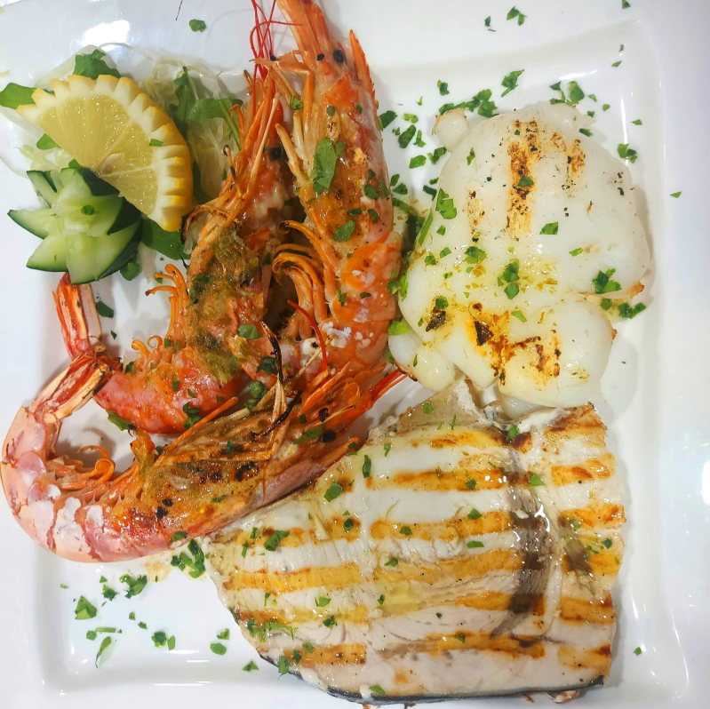 Un piatto bianco con sopra pesce alla griglia, gamberi e cetrioli