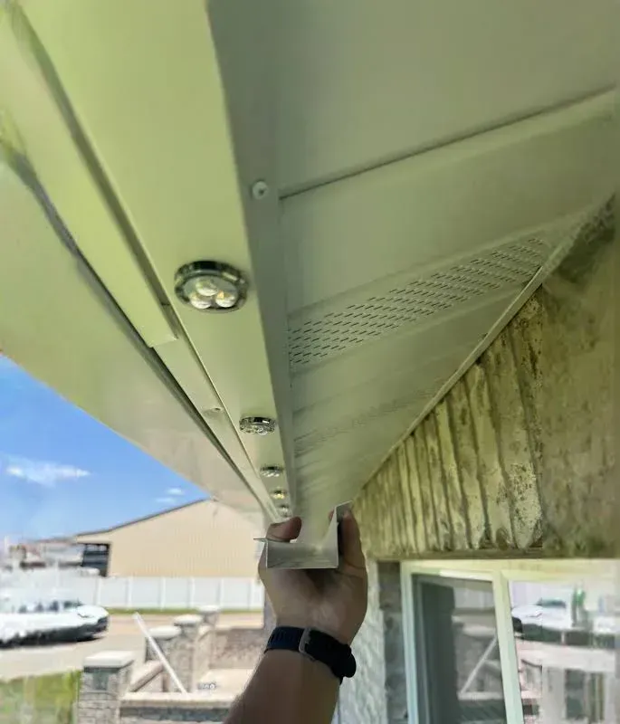 Hand securing a metal soffit panel under a roof eave beside a window and stone wall.