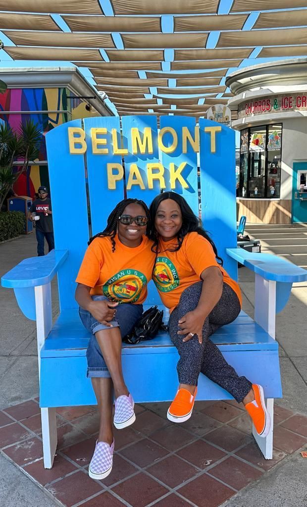 Two women are sitting on a blue chair in front of a sign that says belmont park.