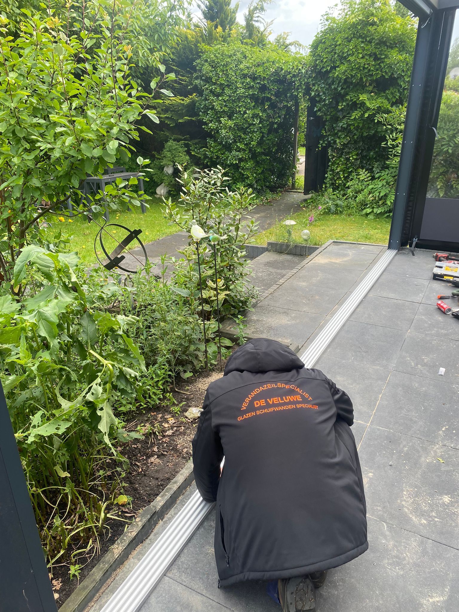 Een man in een zwart jasje knielt op de grond in een tuin.