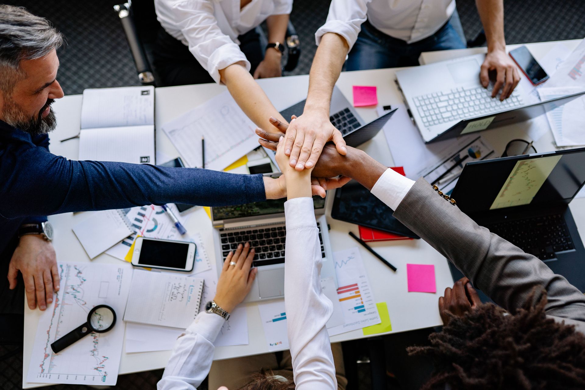 Team hands stacked together over a meeting table with laptops, papers, and sticky notes