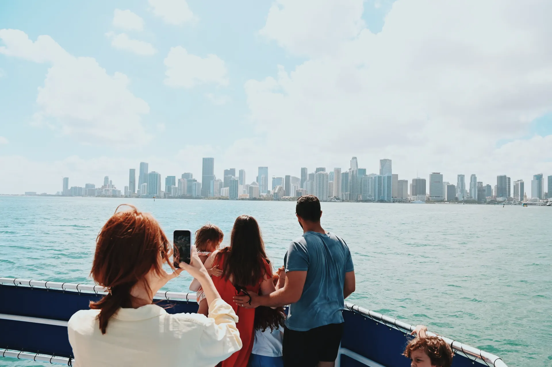 Visitors standing on the upper deck of a sightseeing boat while photographing the Miami skyline across Biscayne Bay on a clear afternoon.