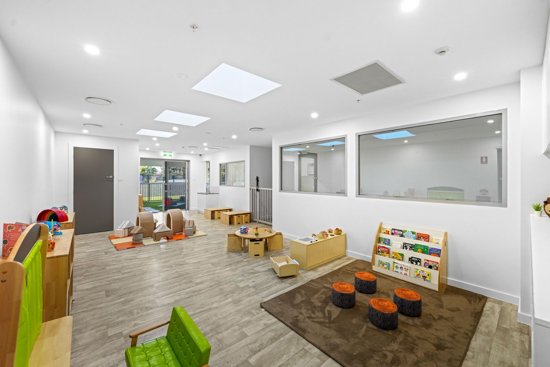 Childcare room with wooden furniture, books, and play area, featuring a brown rug and windows.