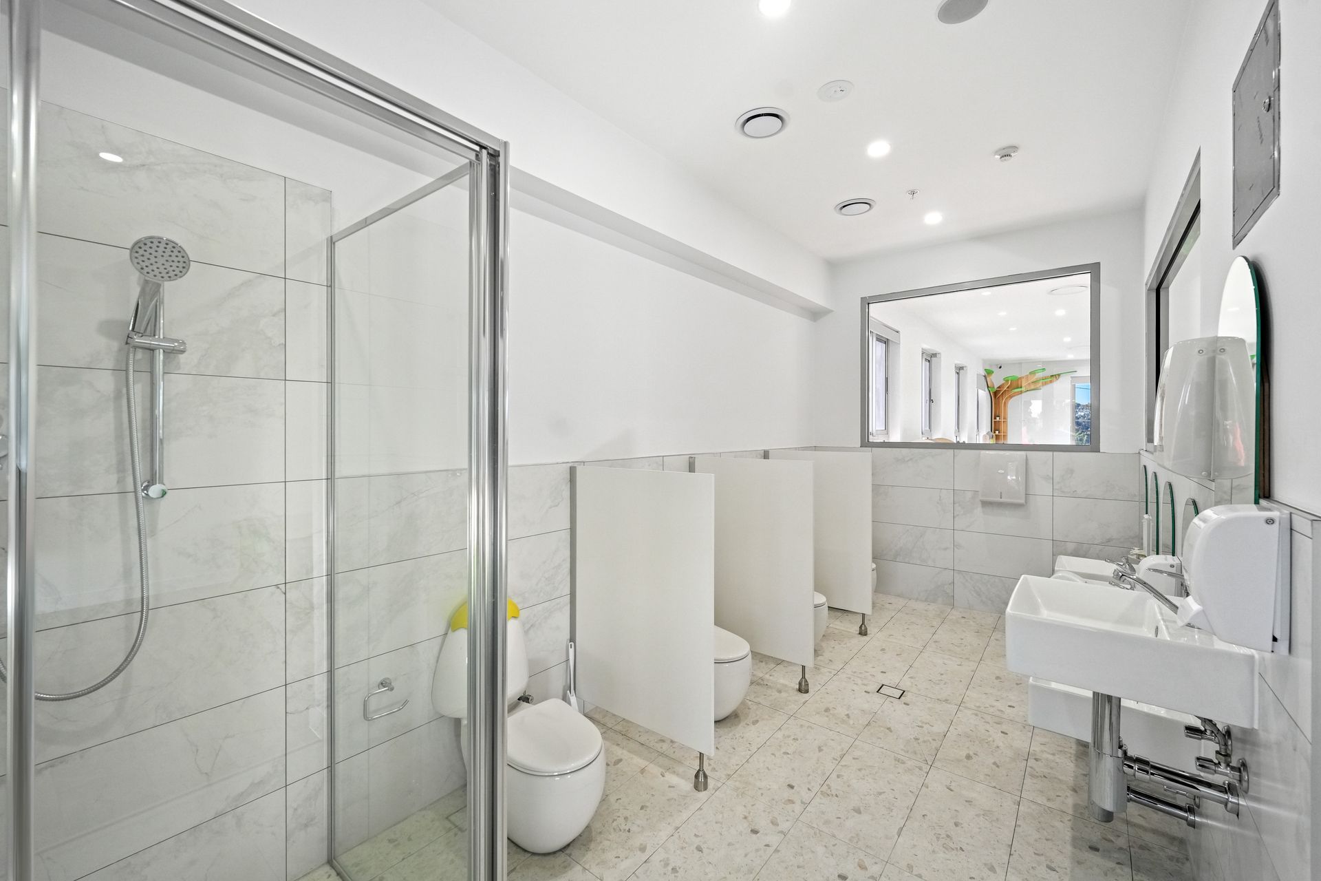 Children toilets with shower stall, white walls, toilets, sinks, and large mirror.