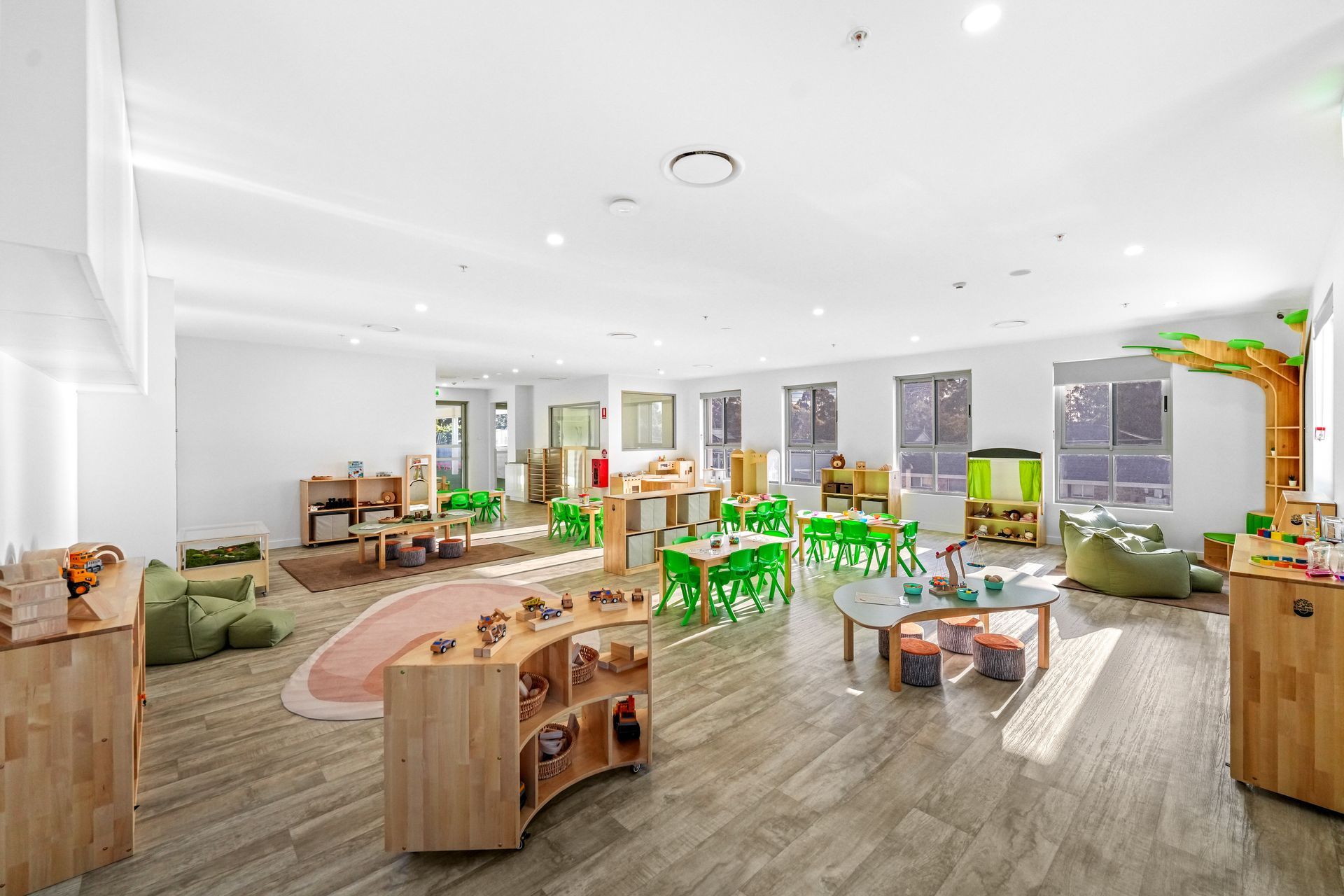 Bright, open-plan preschool classroom with wooden furniture, green chairs, and a play area.
