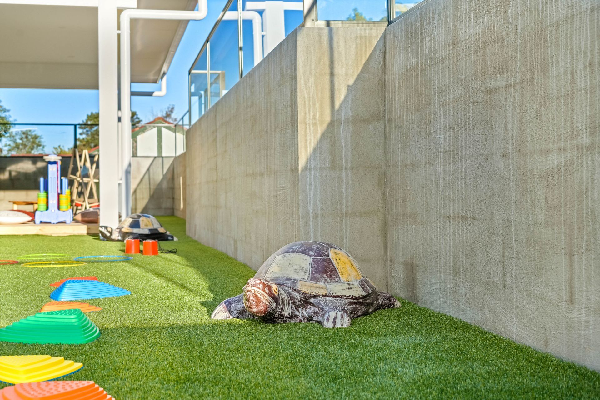 Outdoor play area with a large turtle statue on artificial grass. Concrete wall and colorful play equipment are visible.
