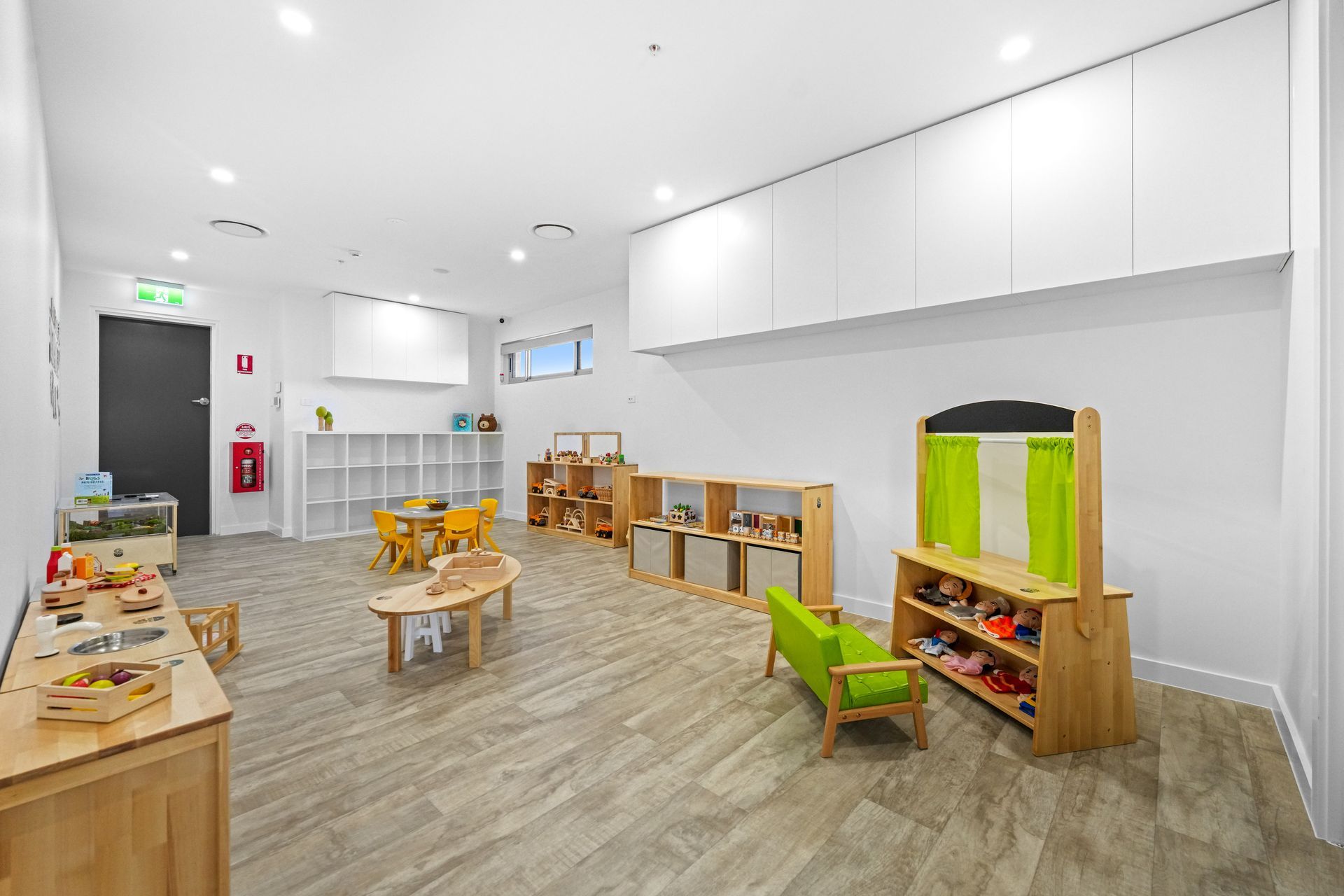 Childcare classroom with wooden furniture, toys, and play area on wood-look floor.