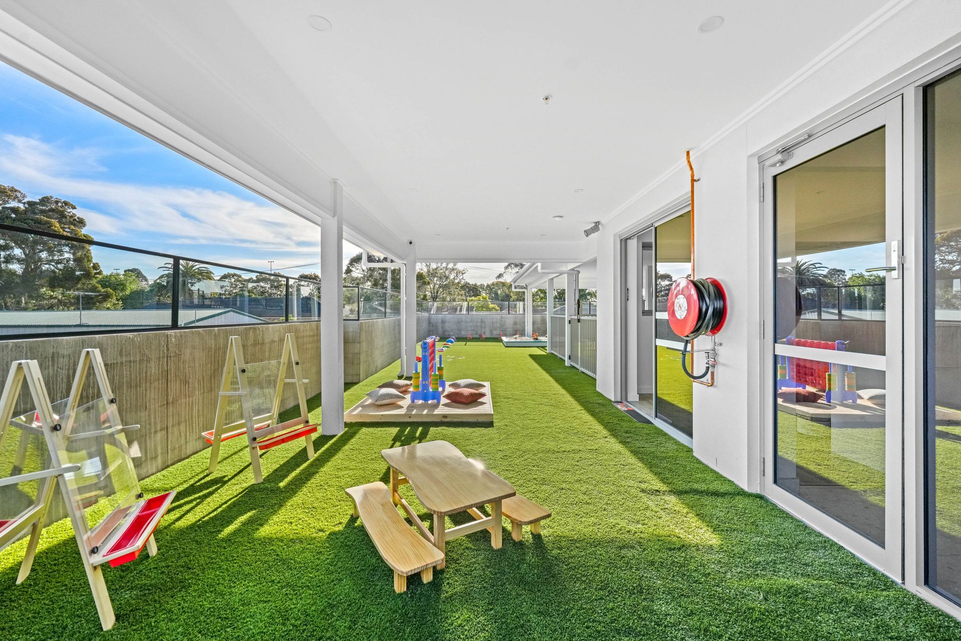 Covered outdoor play area with artificial grass, play equipment, picnic table, and glass doors.