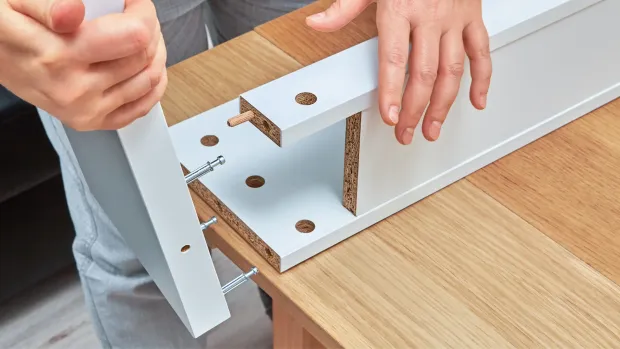 Hands assembling white furniture pieces on a wooden surface, using screws and dowels.
