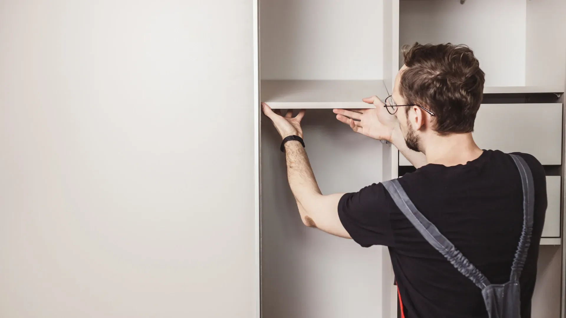 Man installing a shelf in a white closet, wearing glasses and overalls.