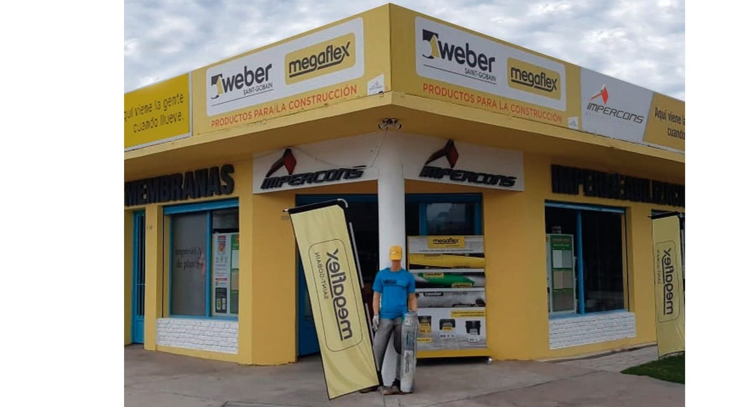 Un hombre está de pie frente a un edificio amarillo con un cartel. El edificio tiene letreros de materiales de construcción.