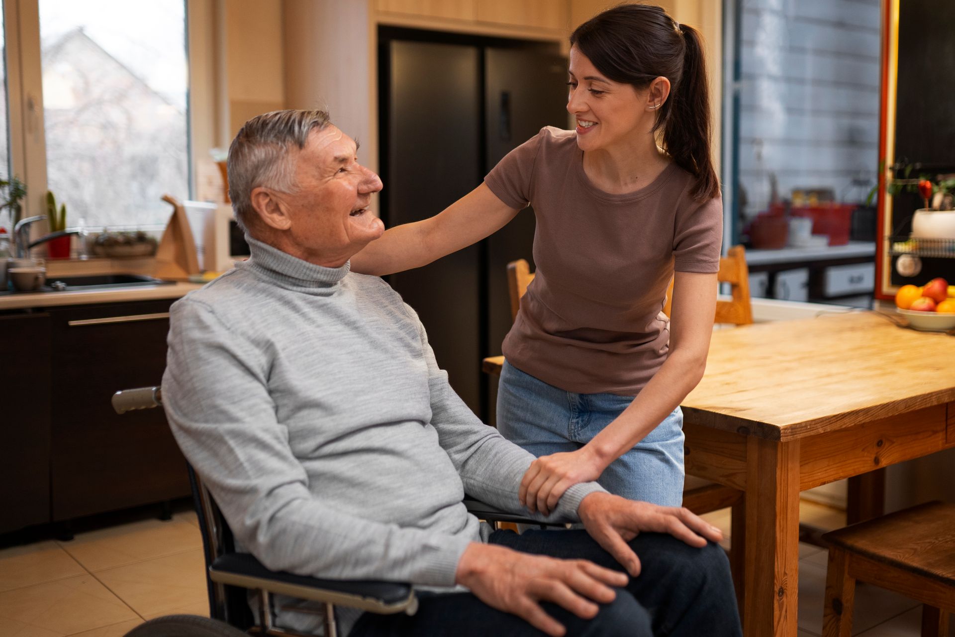Young woman and elderly man in a wheelchair smiling at each other in a kitchen
