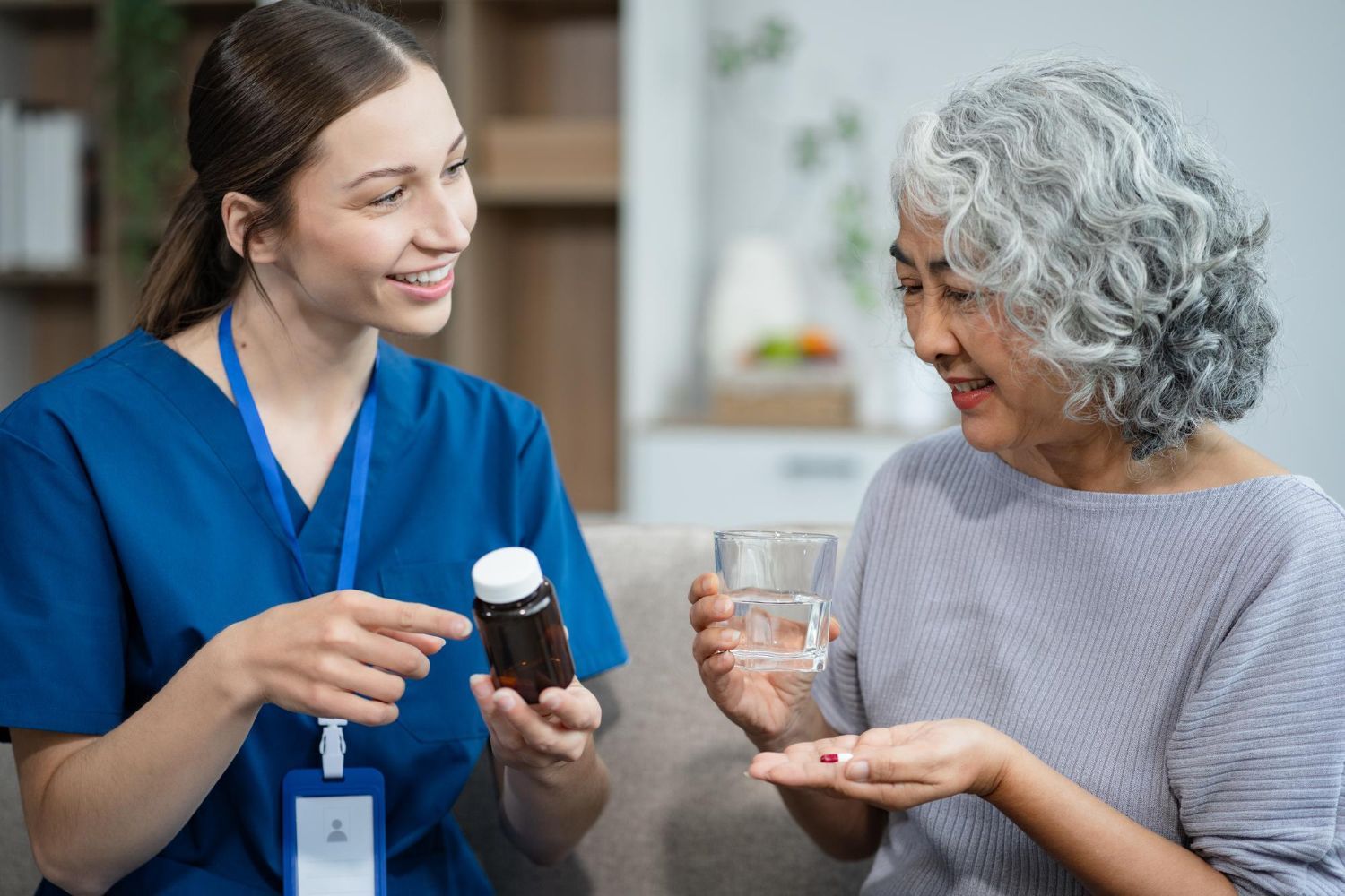 Caregiver in blue scrubs shows medicine bottle to smiling woman holding a glass of water