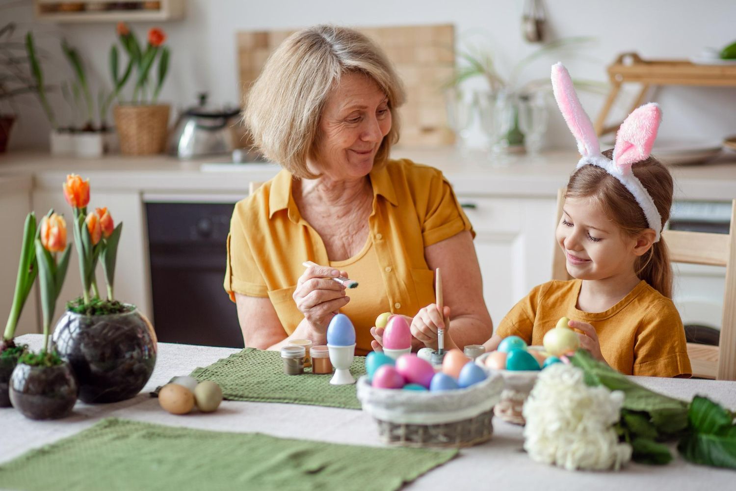 Woman and child painting Easter eggs at a kitchen table. The child wears bunny ears.