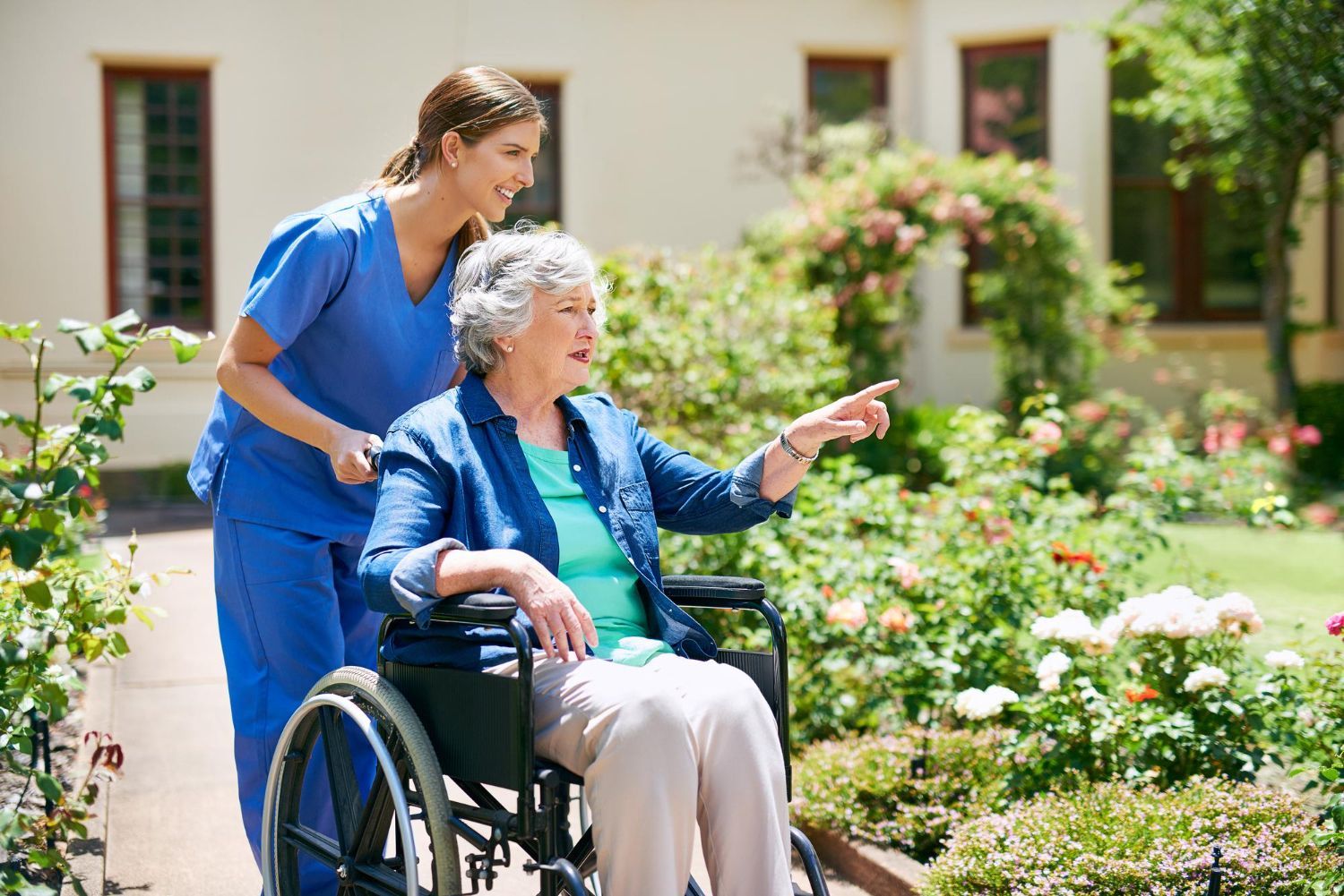 Caregiver in blue assists elderly woman in wheelchair in a flower garden, both looking ahead and pointing.