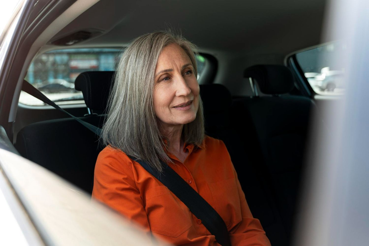 Woman with gray hair wearing an orange shirt seated in the back of a car, looking out the window, seatbelt fastened.