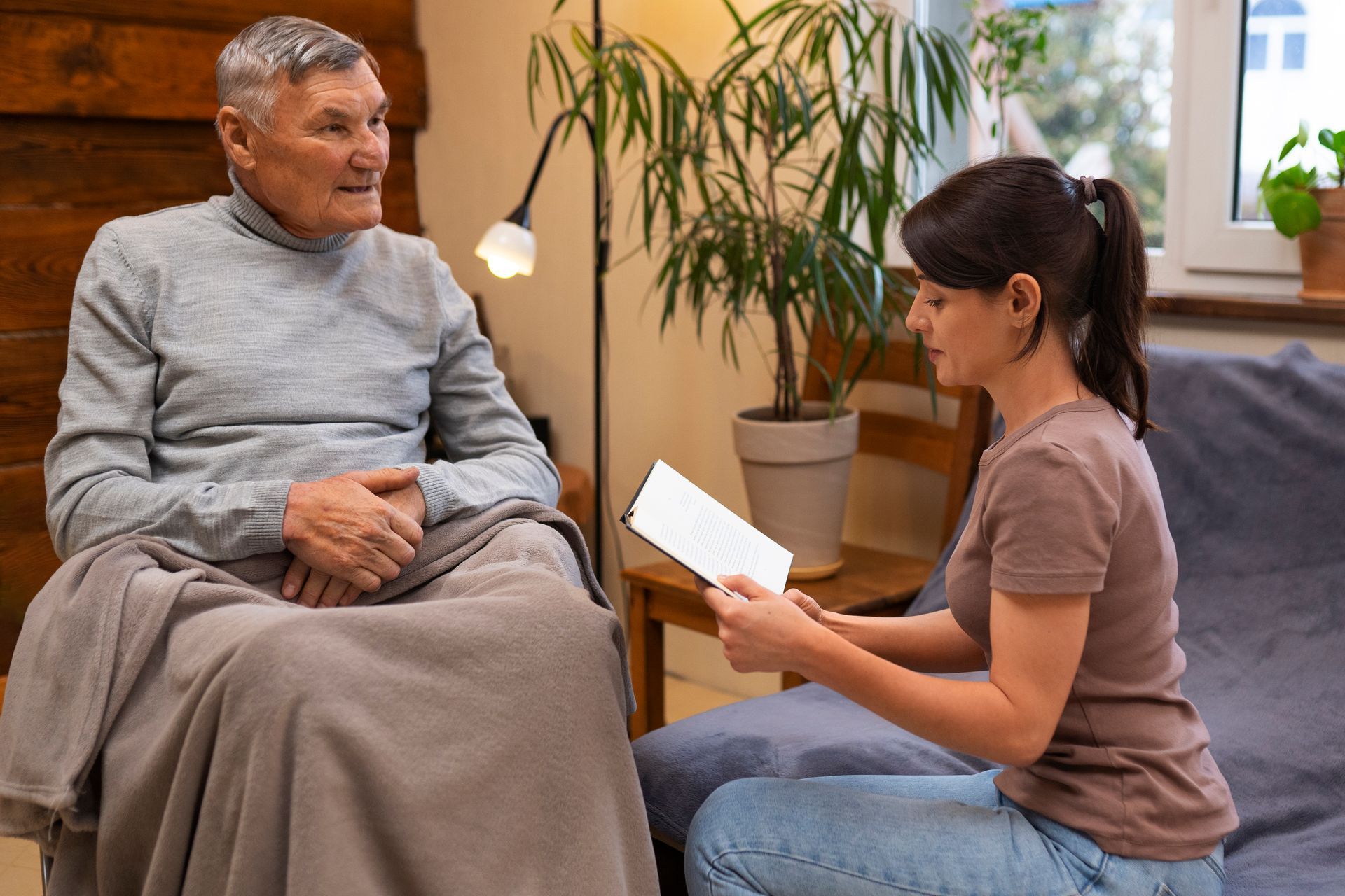 A woman is reading a book to an elderly man who is sitting on a couch.