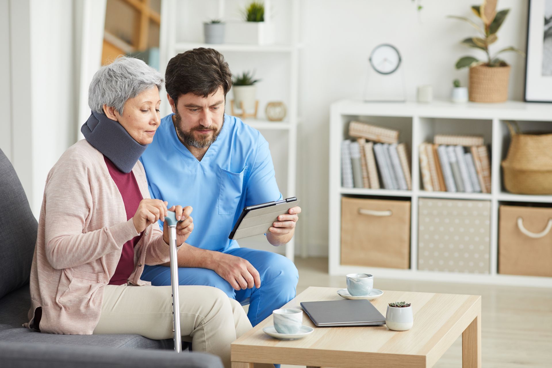 A healthcare worker showing a tablet to a patient with a neck brace, seated, using a cane, in a home setting.