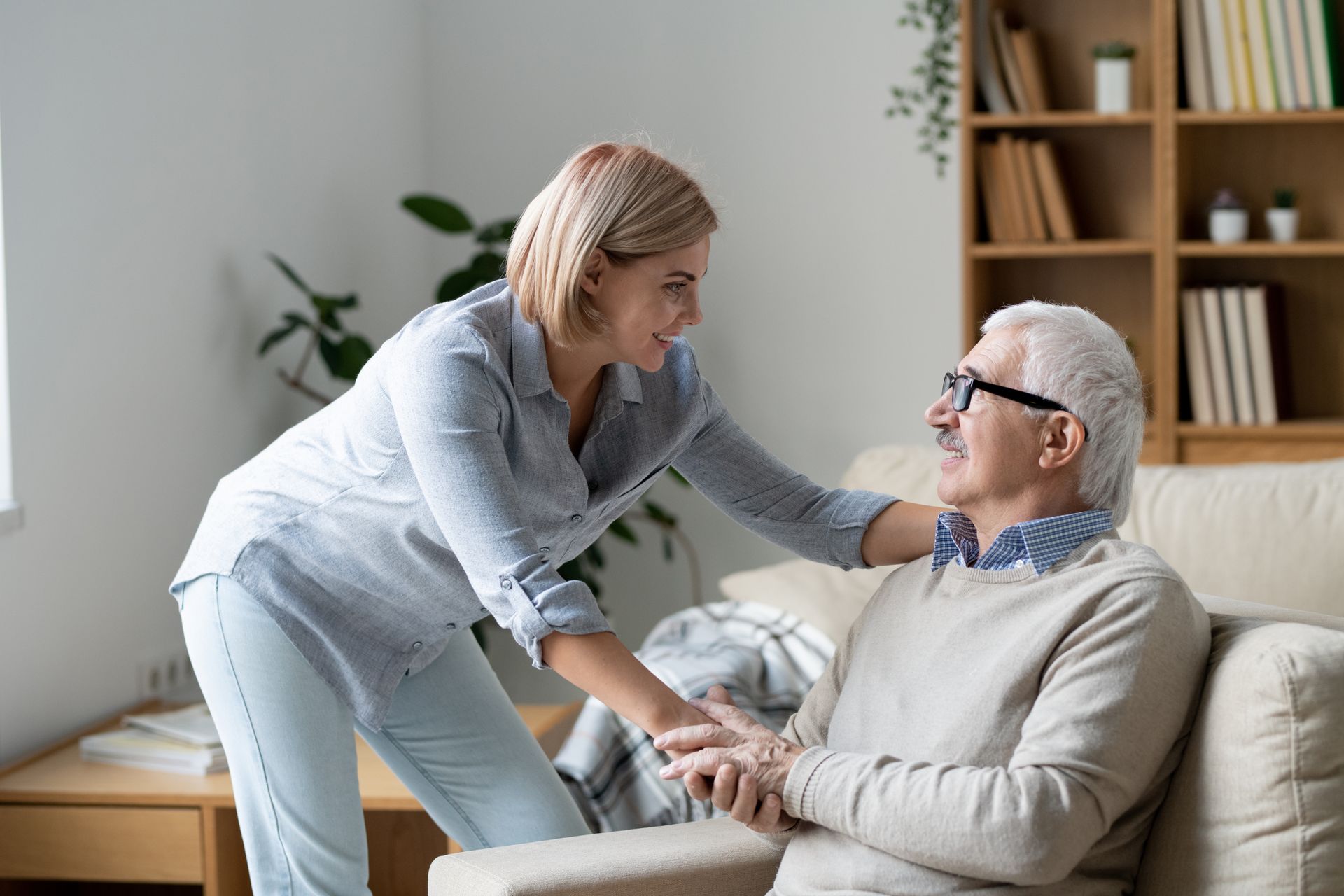 A woman is standing next to an older man sitting on a couch.
