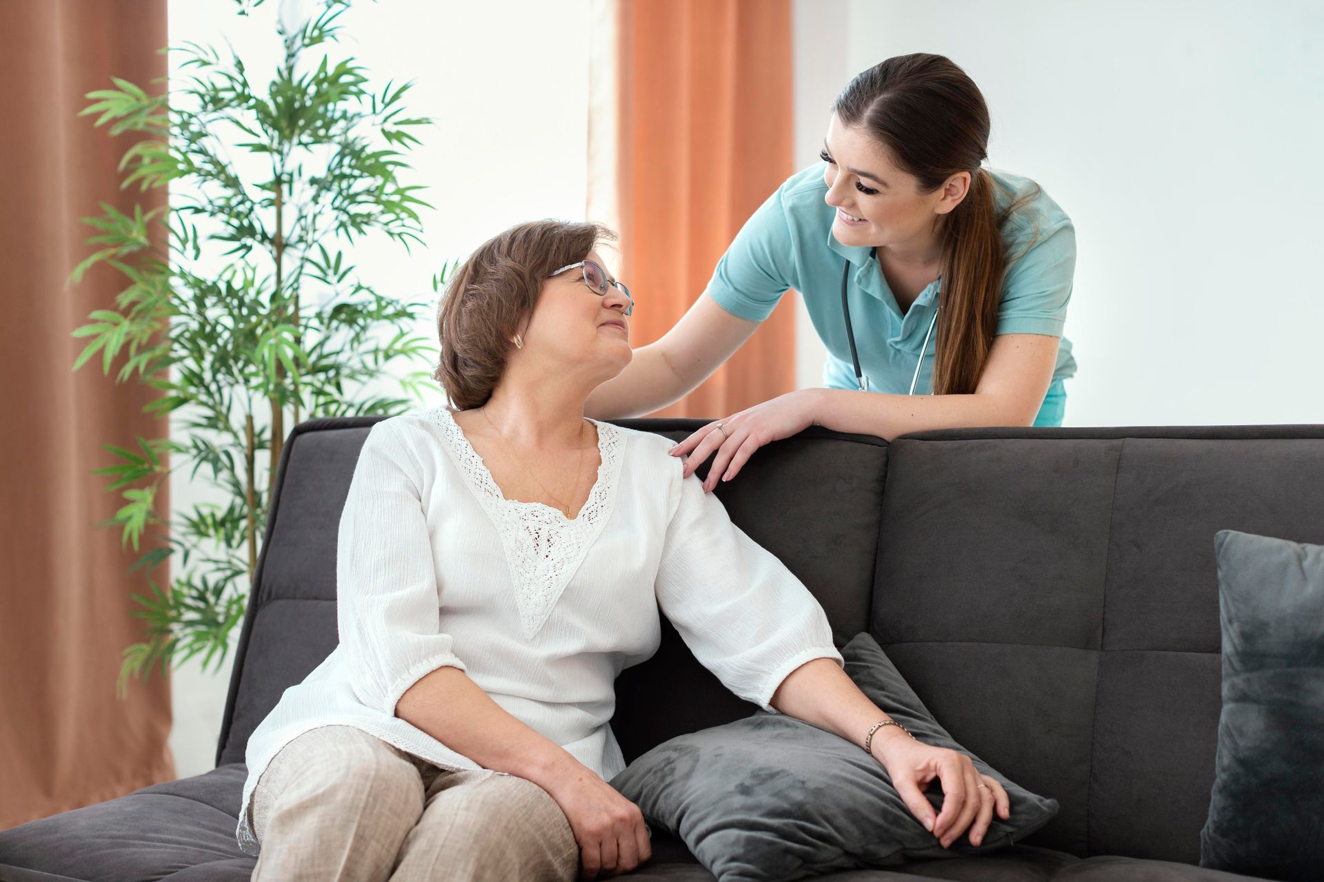 Woman on couch with younger person leaning on couch, smiling. Living room setting.