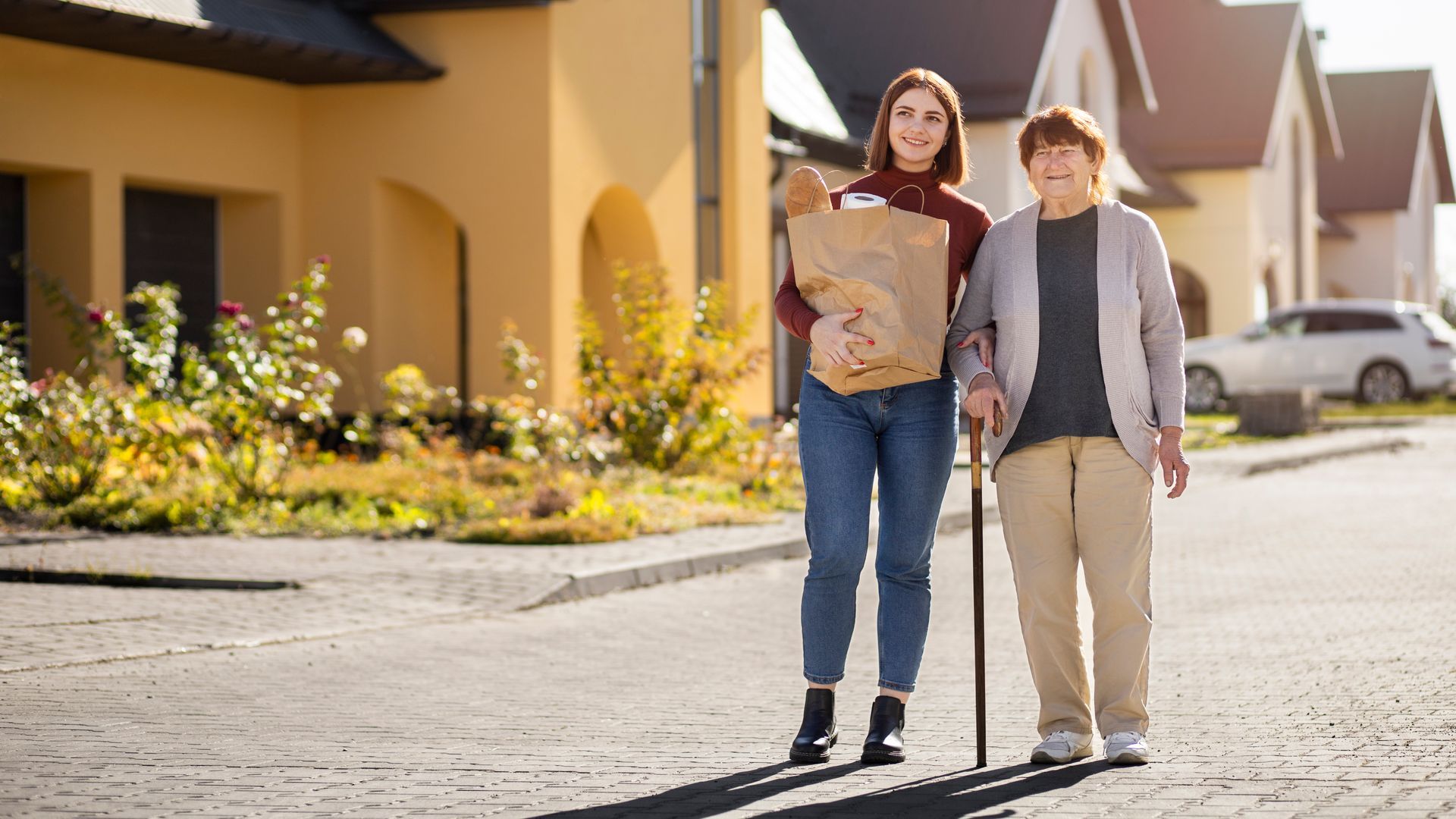 Woman assisting older person walking outside, carrying groceries.