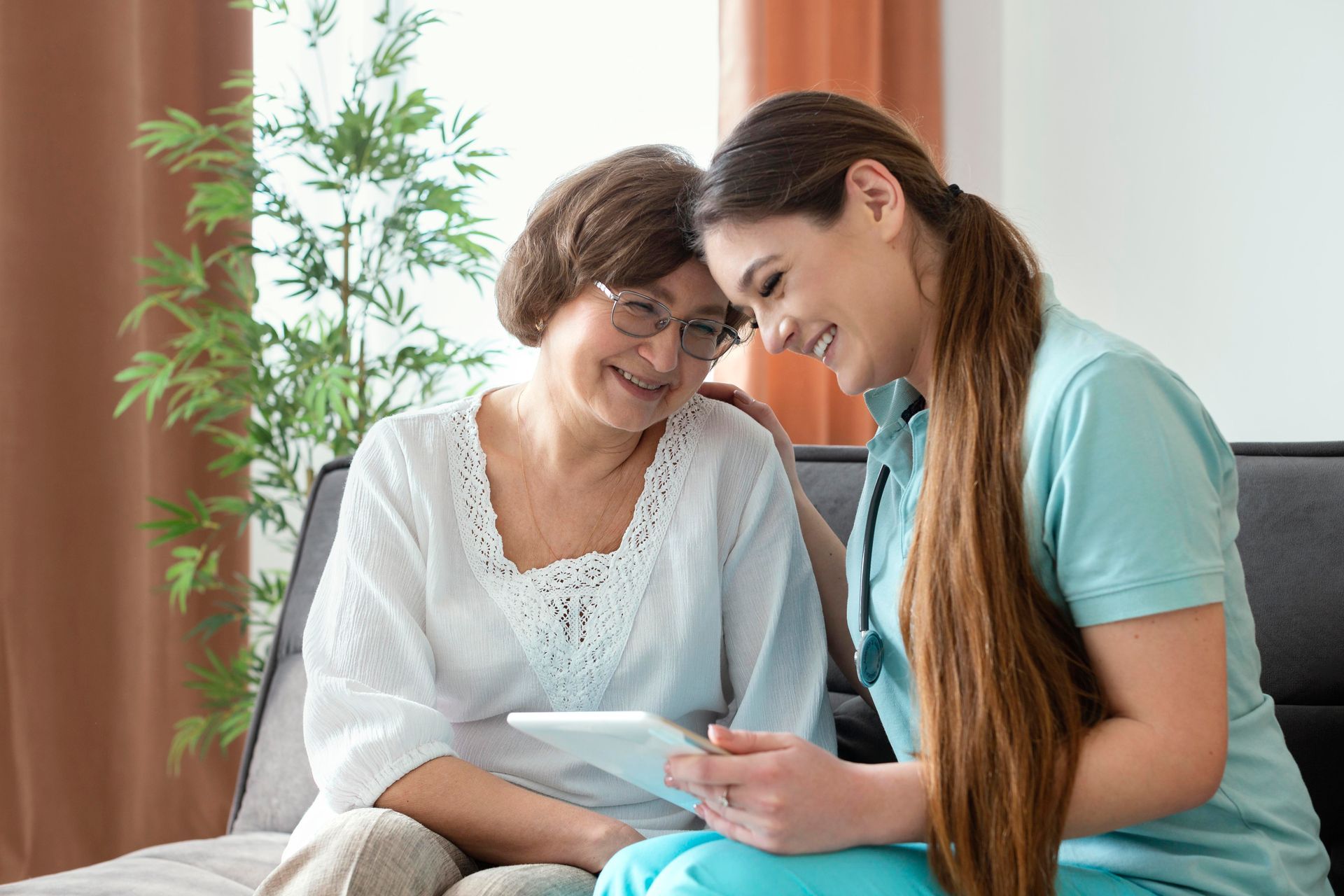 Woman with glasses and caregiver looking at tablet, smiling indoors.