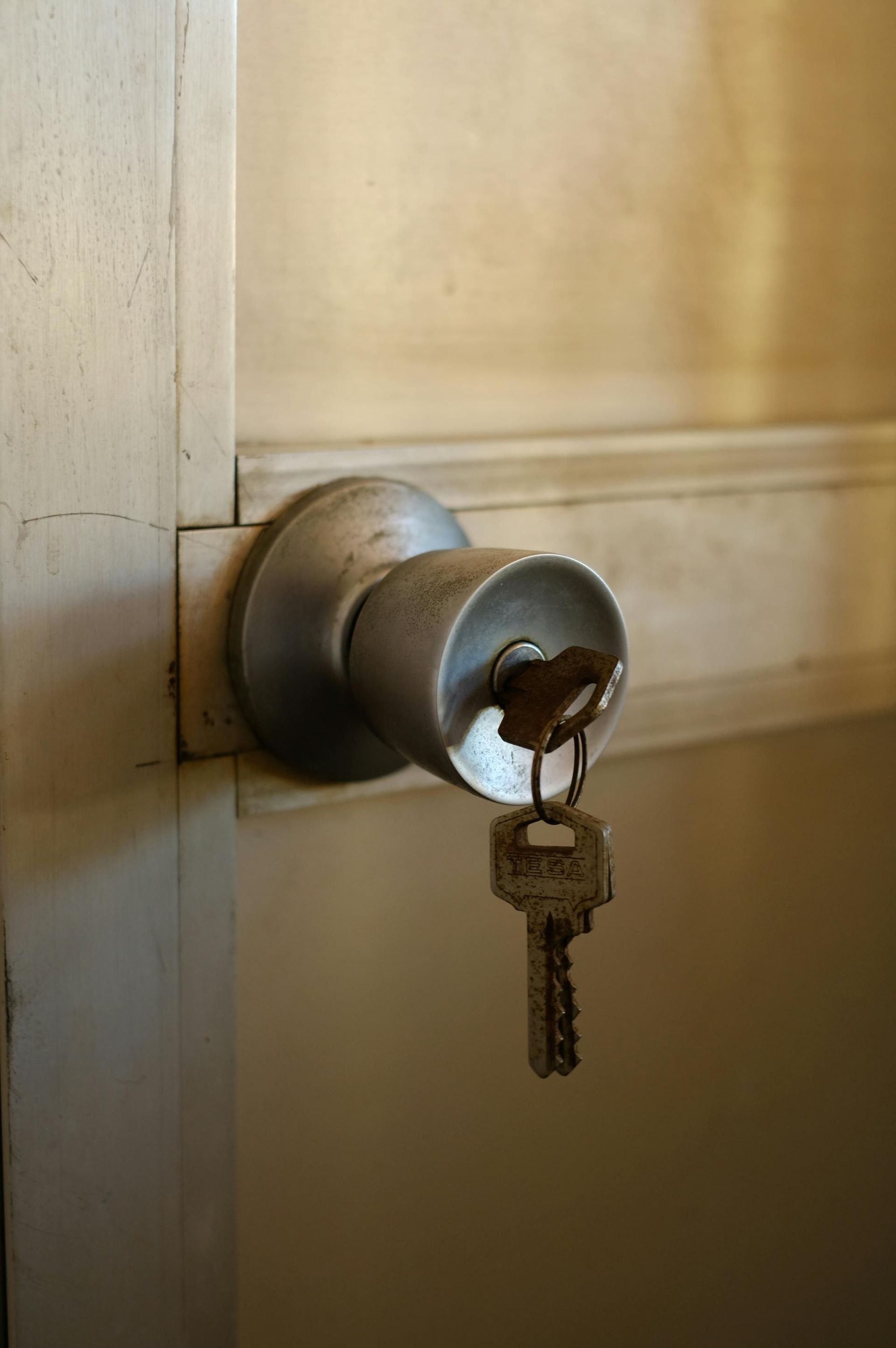 A silver metal doorknob attached to a light-colored wooden door, with a brass key hanging from the lock.