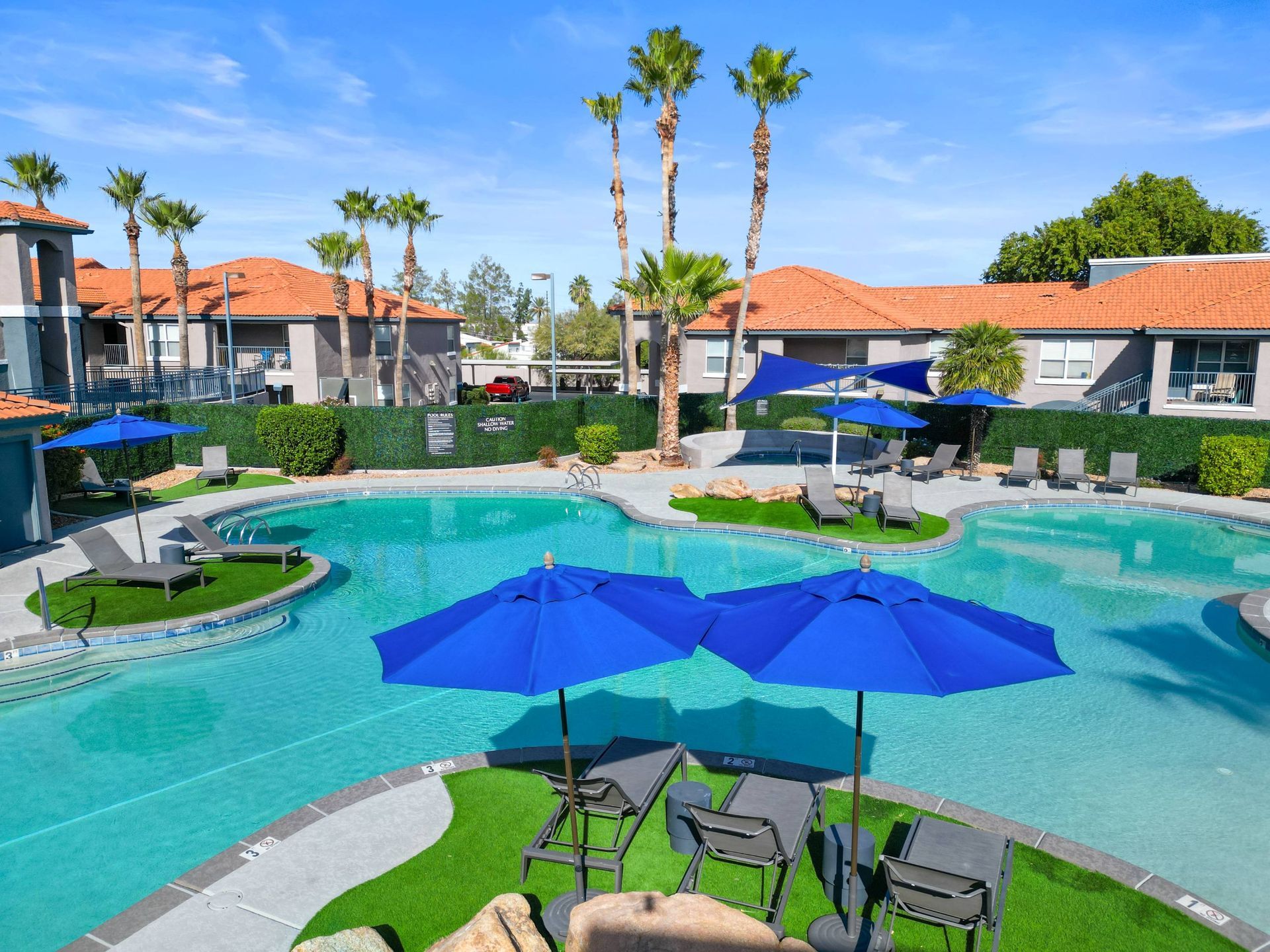 Outdoor pool area at a multifamily community with blue umbrellas, lounge chairs, and palm trees.